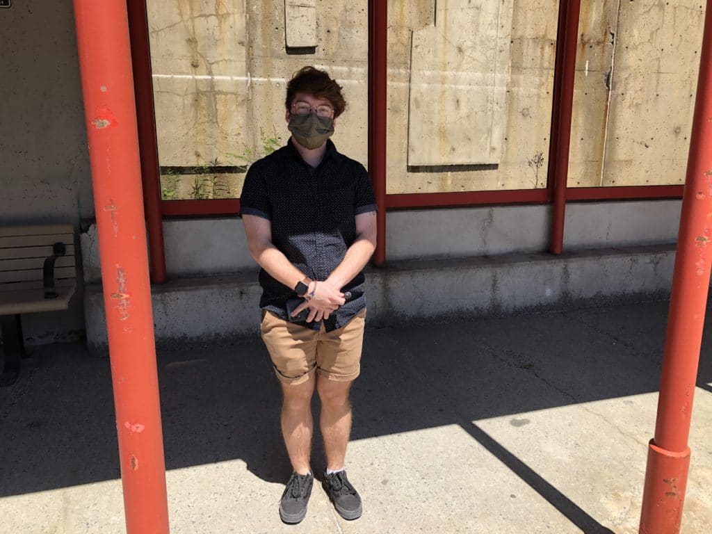 Photo of Matthew Gradelle, wearing a cloth face mask, arms crossed in front of his body, waiting for the bus.