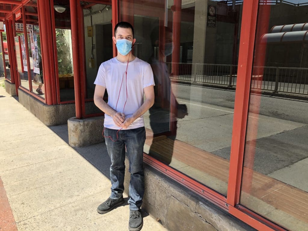 Photo of Patrick Grimsley, waiting for a bus, wearing a face mask with one headphone in