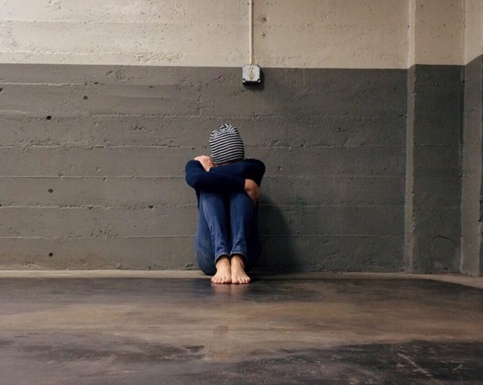 A person sits alone with their back against a wall, their head covered and pulled down to their knees