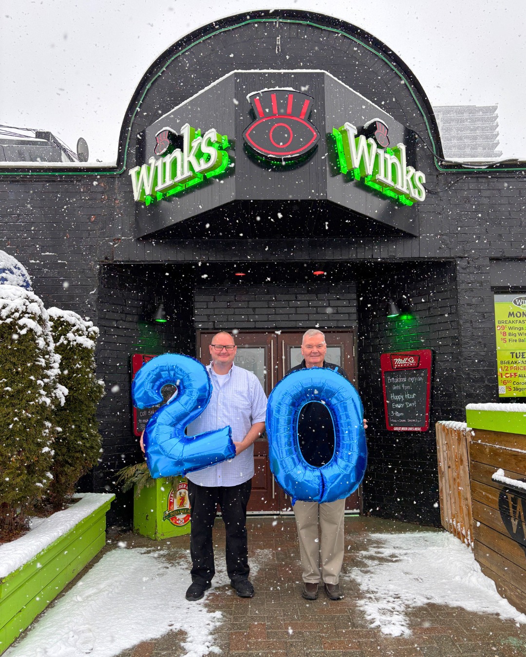 Adam Winkler wears glasses, a button down shirt and black pants and shoes. He's holding a blue balloon shaped like the number two. His father Dennis wears a black jacket, beige khakis and brown shoes. He holds a blue balloon shaped like the number 0. They stand smiling in front of Winks Eatery while snow falls.